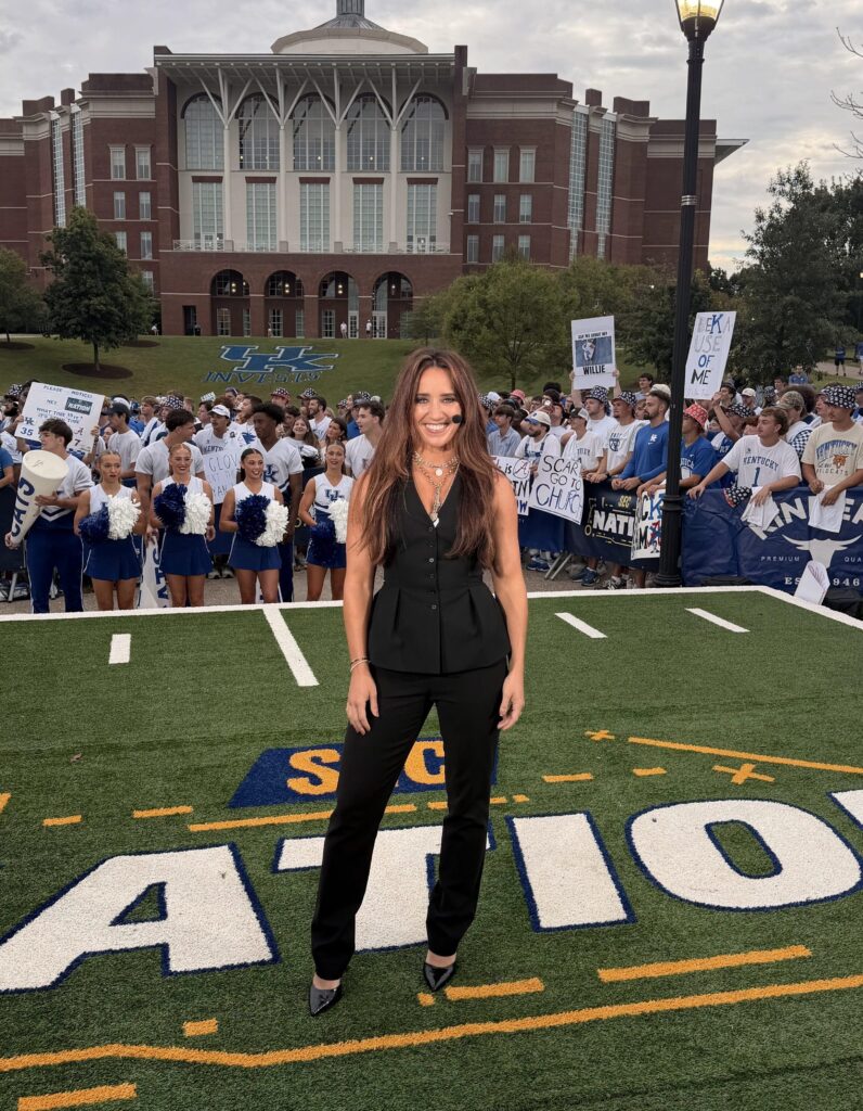 A woman wearing a black outfit stands on the sidelines of a football field. Behind her are several fans and cheerleaders wearing blue and white.