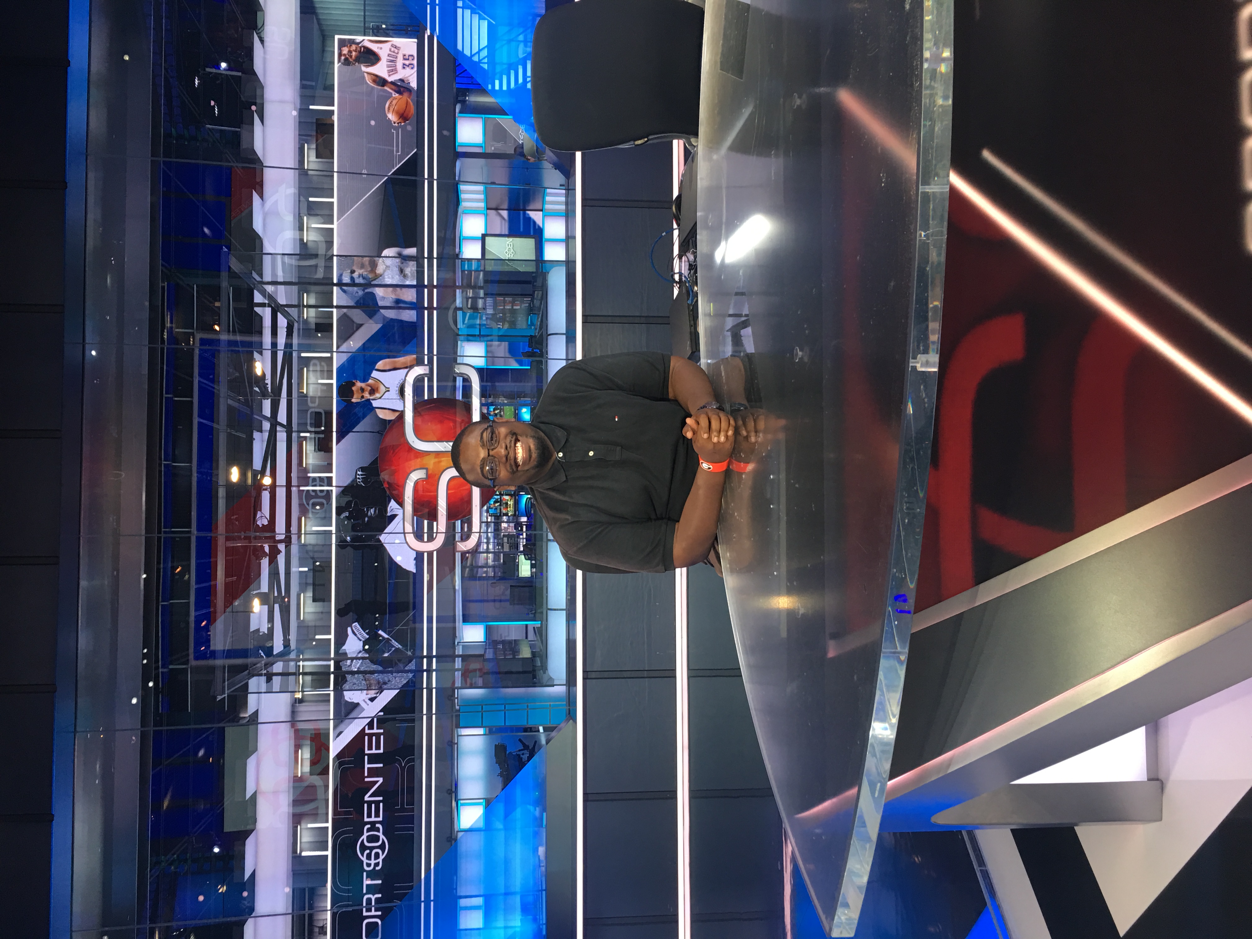 A young man in a black polo sis. at a large glass table on set in a sports production studio.