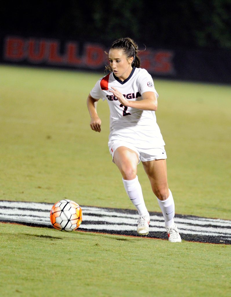 A woman in a white soccer uniform runs to kick a ball on a soccer field.