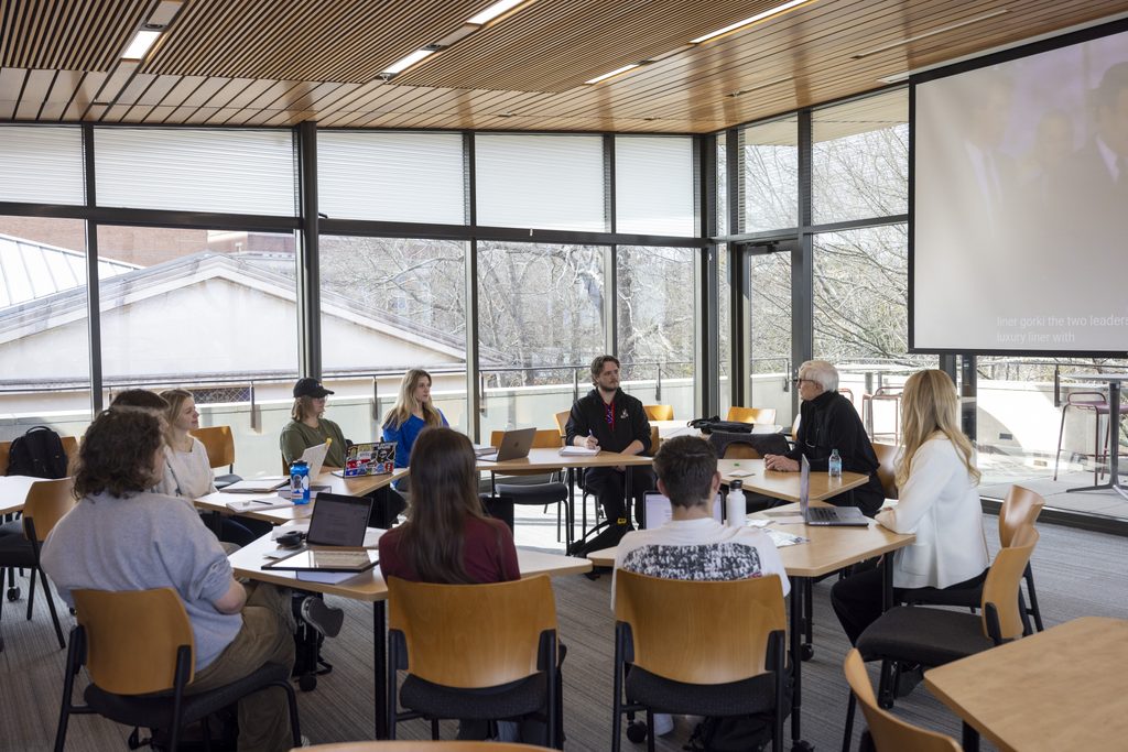 An older man speaking to a group of students seated at tables in a classroom with floor-to-ceiling windows.
