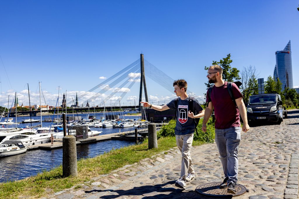 Two young men walking together down a cobblestone street alongside a river marina with sailboats.