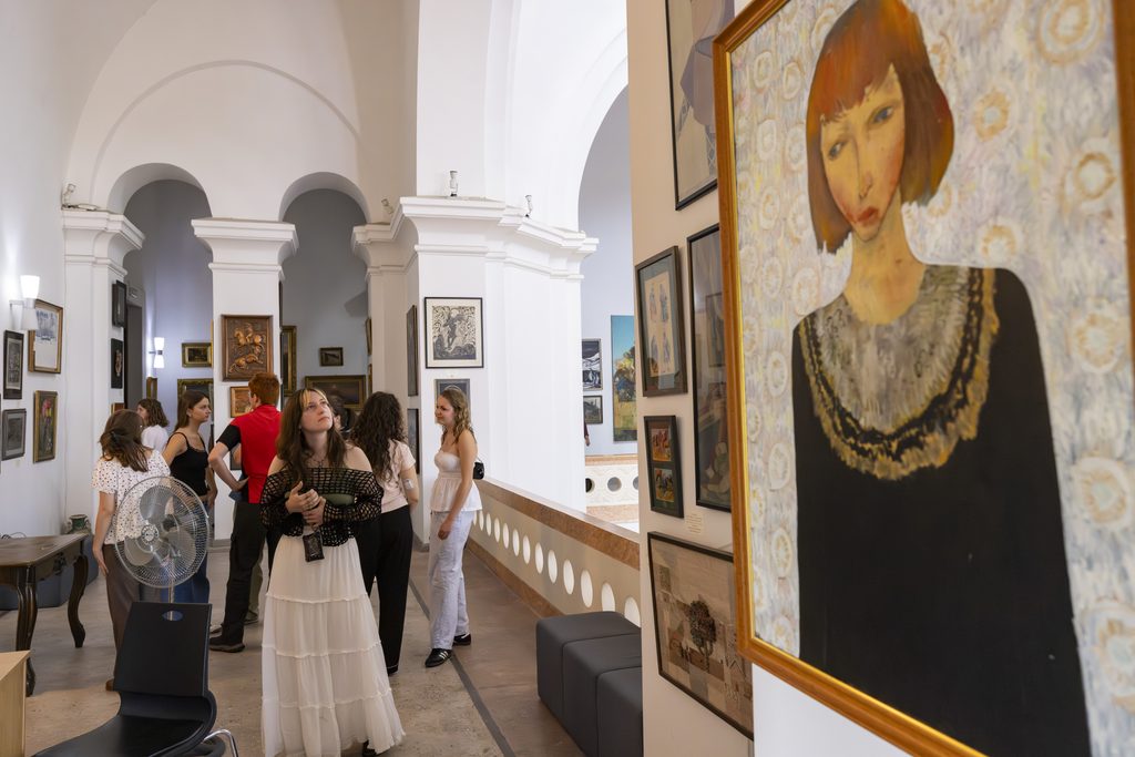 Two students standing in a gallery looking at classic portraits hanging on a neutral wall.