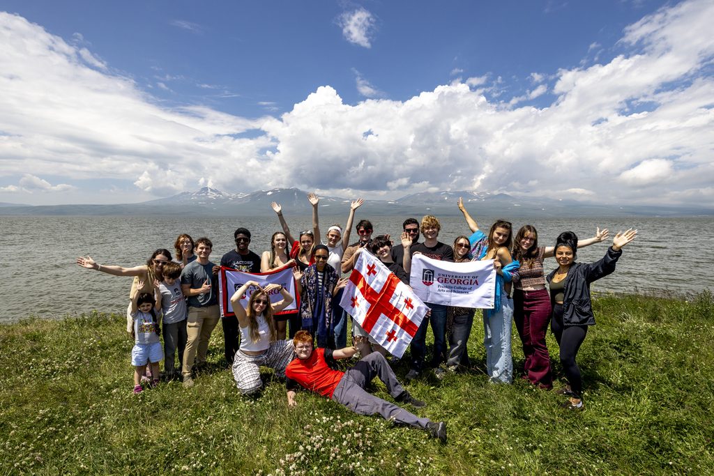 A group of people posing together with flags on a grassy meadow overlooking a wide, calm lake.