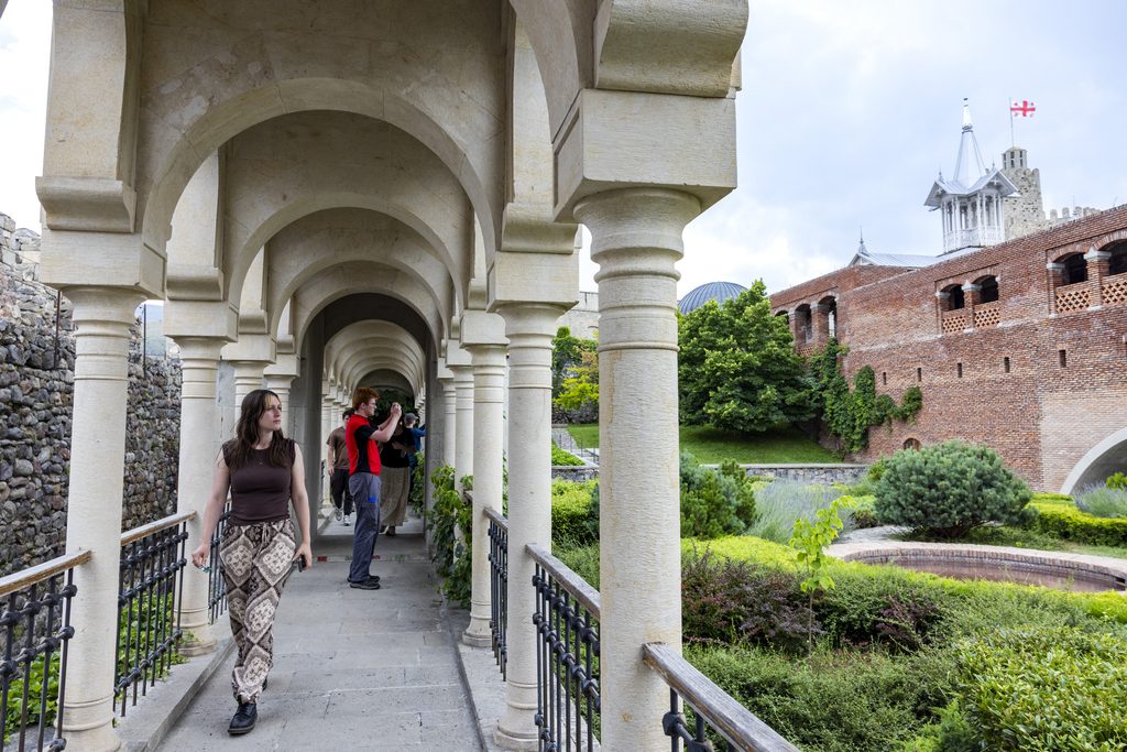 A young woman walks along a narrow, columned walkway flanked by a garden and a brick and stone fortress.